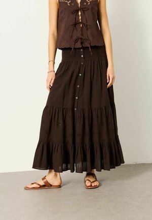 Mujer con top sin mangas marrón oscuro bordado con lazos y falda larga a capas a juego con botones en la parte frontal, acompañada de sandalias.