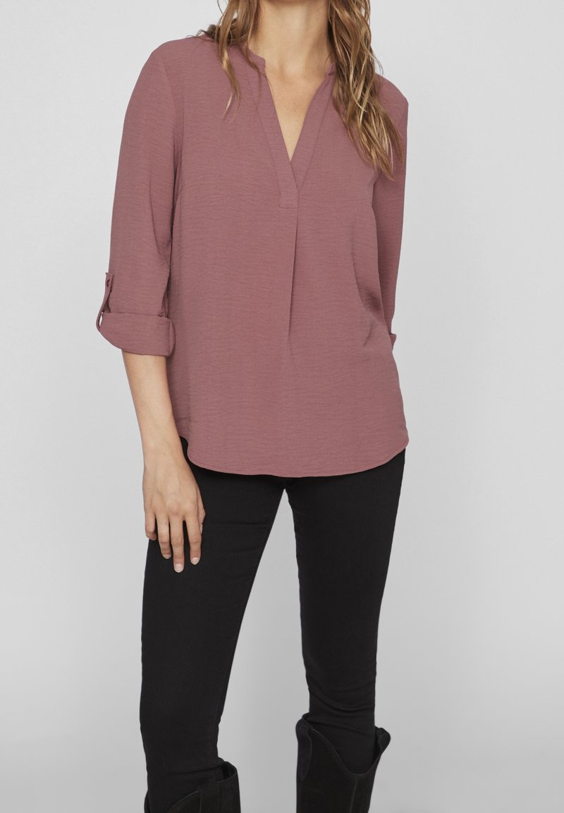 Femme portant un chemisier rose poudré à manches longues avec des poignets retroussés et un pantalon noir sur un fond gris clair uni.