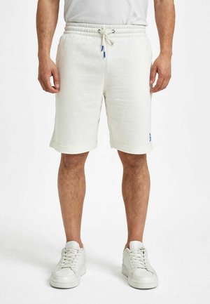 Uomo che indossa pantaloncini bianchi con coulisse, scarpe da ginnastica bianche e una camicia bianca, in piedi contro uno sfondo bianco uniforme.