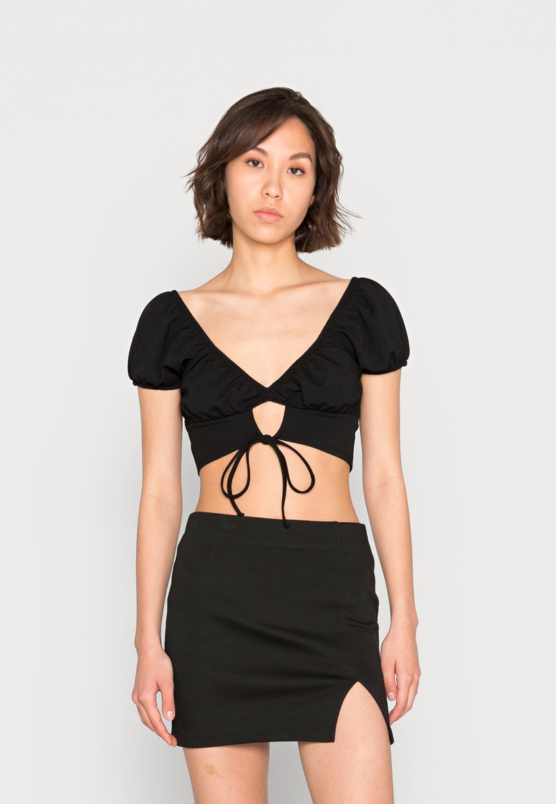 Top curto preto com mangas bufantes, decote V e detalhe de laço na frente. Combinado com uma saia mini preta de cintura alta com uma fenda lateral.