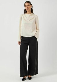 Lys beige langærmet top med en tekstureret overflade, parret med vide sorte bukser. Modellen har spidse sorte stiletter på. Simpelt, elegant design.
