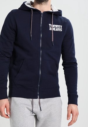 Felpa con zip di colore blu navy, con cappuccio foderato in grigio, logo bianco sul petto e lacci a contrasto. Presenta tasche frontali e polsini a costine.
