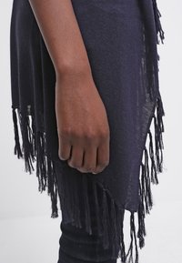 Námořnicky modrý šalen s třásněmi z pletené látky, s dlouhými střapci podél okraje, přehozený přes tmavé látkové pozadí.