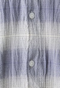 Košile na knoflíky s texturovaným pruhovaným vzorem v odstínech modré a bílé, s kulatým bílým knoflíkem na středu.