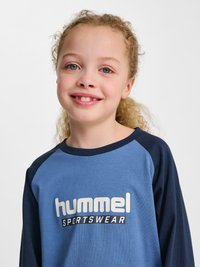 Jeune fille souriante aux cheveux blonds bouclés portant un maillot de sport Hummel bleu et marine sur un fond uni.