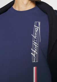 Námořnické bavlněné tričko s bílou podpisovou grafikou a vertikálními liniemi, s barevným pruhovaným detailem na spodním okraji.