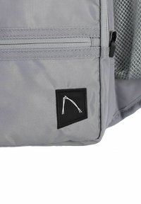 Sivi platneni ruksak s teksturiranim površinom, s crnom zakrpom sa bijelim logom, i patentnim zatvaračem na vrhu.