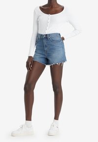 Haut à manches longues blanc à côtes avec détails de boutons, associé à un short en denim bleu taille haute avec des ourlets effilochés. Des baskets blanches complètent le look.