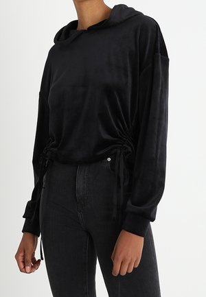 Musta pitkähihaisen hupullisen paidan, jossa on rypytetyt kiristysnauhavivut sivuilla, yhdistetty korkeavyötäröisiin mustiin farkkuihin neutraalilla taustalla.