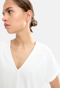 Hvid, kortærmet bluse med en dyb V-hals. Blødt stof med en glat tekstur. Simpelt design, minimal sømdetaljer og ingen mønstre.