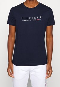 Námořnická modrá bavlněná tričko s kulatým výstřihem a krátkými rukávy, s bílým, červeným a modrým textem "HILFIGER NEW YORK".