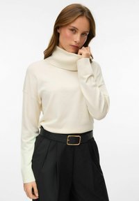 Kremowy sweter z golfem w luźnym kroju, z długimi rękawami i zaokrąglonym dekoltem, noszony z czarnymi spodniami o wysokim stanie i czarnym paskiem.