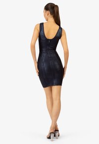 Robe ajustée marine avec un décolleté plongeant dans le dos, motif texturé et fermeture éclair à l'arrière, associée à des sandales à talons transparentes.