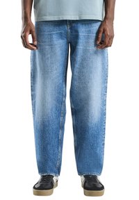 Jasnoniebieskie dżinsy o luźnym kroju, z widocznym blaknięciem i detalami przypominającymi fałdy. Połączone z czarnymi trampkami i jasną koszulą.