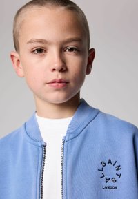 Cazadora bomber azul claro con cuello acanalado, cremallera en la parte frontal y detalle de logo bordado en negro. Camiseta blanca visible debajo. Textura suave.