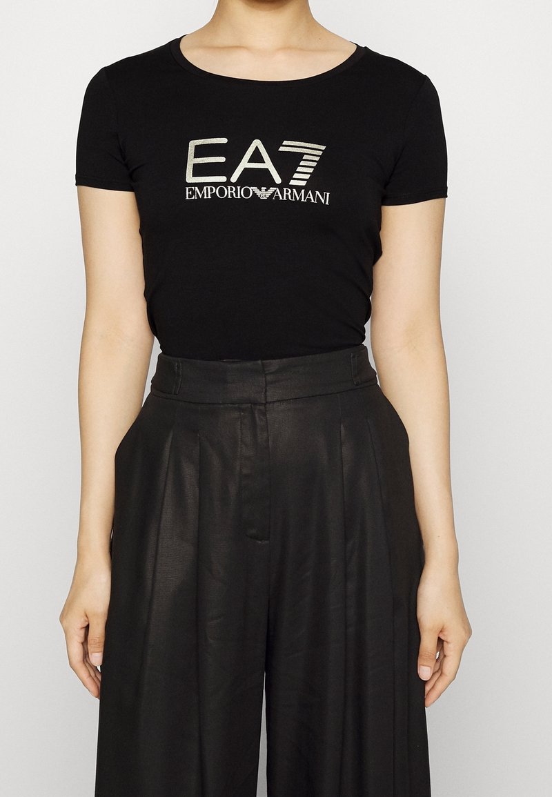 Camiseta negra de manga corta con escote en pico, que presenta un logo metálico "EA7 EMPORIO ARMANI" en el centro, combinada con pantalones negros de talle alto.