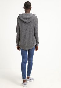 Grijze gebreide hoodie met een achternaad en een capuchon, gecombineerd met blauwe denim jeans en grijze sneakers. De stof heeft een gestructureerde, geribbelde uitstraling.