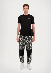 Ung mann iført en svart EA7 t-skjorte, digitale kamuflasjebukser med svarte lapper, hvite joggesko og en svart crossover-bag.