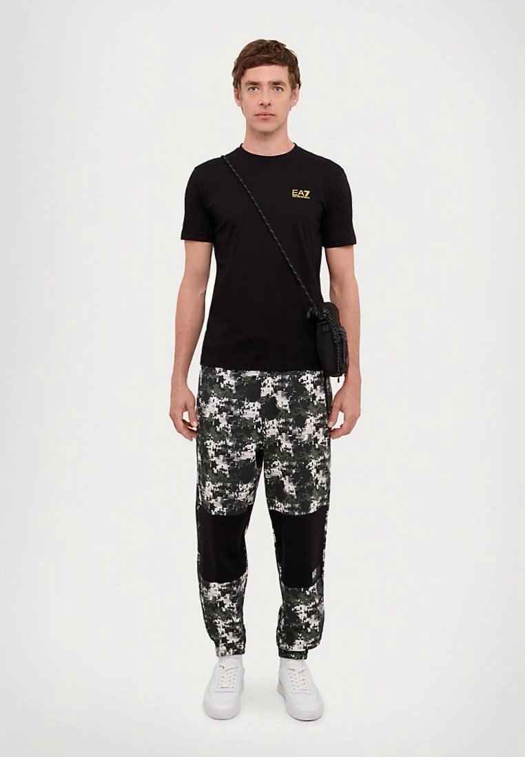 Jeune homme portant un t-shirt noir EA7, un pantalon en camouflage numérique avec des patches noirs, des baskets blanches et un sac bandoulière noir.