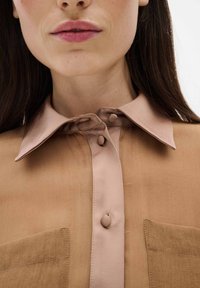 Chemise marron clair avec une texture douce, dotée d'un col contrastant plus clair, de deux poches avant et de boutons ronds.