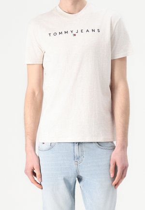 Biała koszulka z krótkim rękawem z napisem „TOMMY JEANS” i logo na piersi, noszona z jasnoniebieskimi dżinsami.