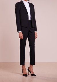 Černý na míru šitý oblek, který zahrnuje přiléhavý blazer se dvěma knoflíky, úzké kalhoty a bílou košili s límečkem. Lesklé černé vysoké podpatky dotvářejí vzhled.