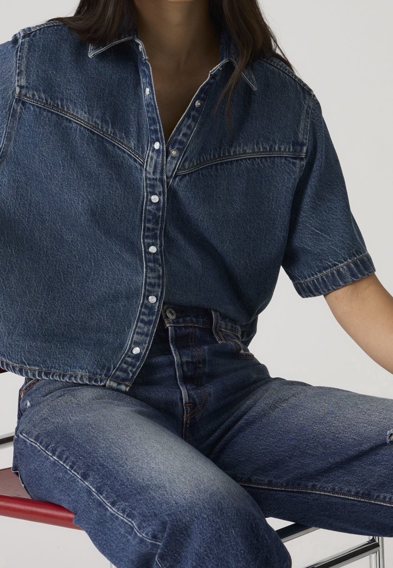 Femme assise portant une chemise en jean bleu foncé à manches courtes et un jean bleu foncé sur un fond uni.