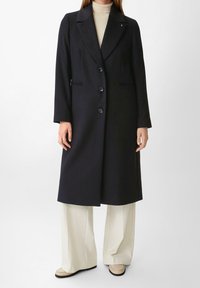 Manteau long marine avec un col à encoches, boutons sur le devant, deux poches latérales, coupe ajustée, associé à un pantalon large crème et des chaussures claires.