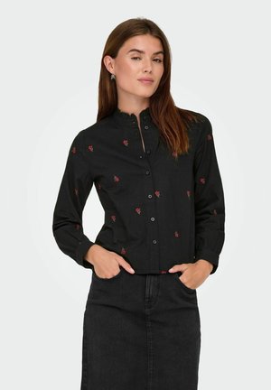 Femme aux longs cheveux bruns portant une chemise noire à boutons avec de petites imprimés de fleurs rouges et une jupe noire, les mains dans les poches.