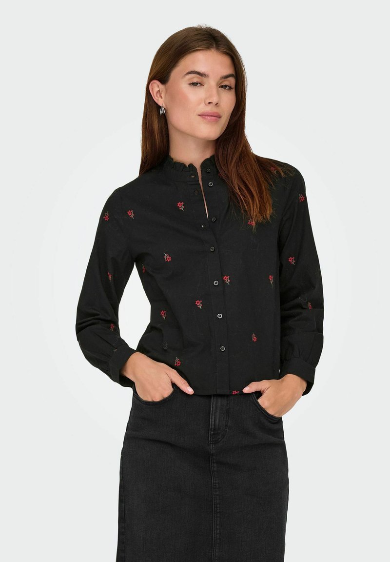 Donna con lunghi capelli castani che indossa una camicia nera con bottoni e piccole stampe di fiori rossi, e una gonna nera, con le mani nelle tasche.