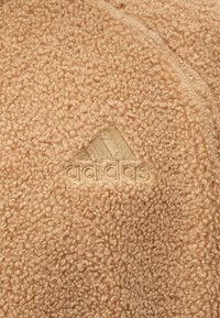 Beige, textúrált fleece anyag, hímezett Adidas logóval, amelyen a trèfli minta és a márkanevéhez illő cérna látható.