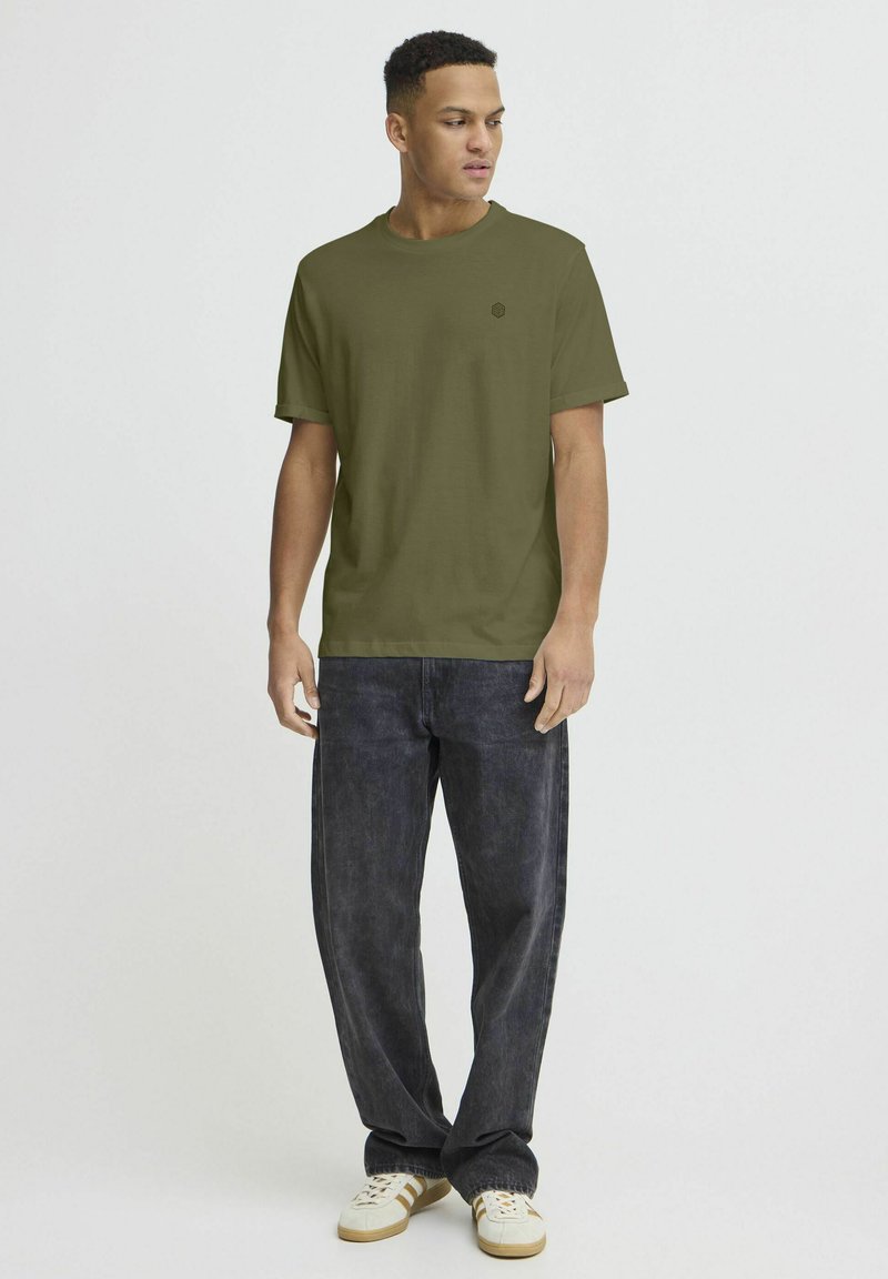 T-shirt verde oliva a maniche corte con scollatura rotonda e un piccolo logo sul petto, abbinato a jeans in denim scuro e sneaker bianche.