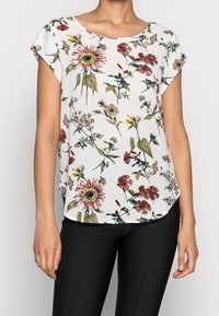Donna che indossa una blusa bianca a maniche corte con stampa floreale multicolore abbinata a pantaloni neri aderenti su uno sfondo semplice.