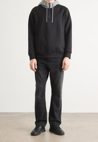 Sudadera negra con capucha gris, bolsillo frontal y cordones, combinada con jeans de lavado oscuro y zapatillas negras sobre un fondo neutro.