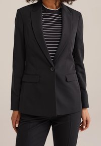 Černý blazer s přiléhavým střihem, s klopami, jednořadovým zapínáním na knoflík a předními kapsami, kombinovaný s pruhovaným topem pod ním.