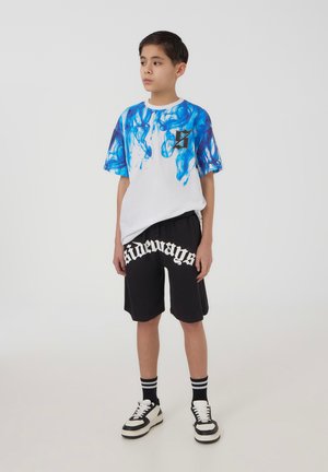 Maglietta bianca con motivo a fiamma blu, pantaloni corti neri con testo bianco "sideways", sneaker in bianco e nero, e calzini a righe bianchi e neri.