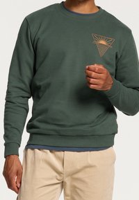 Grön sweatshirt i mjukt tyg med rundad halsringning. Har en triangelgrafik med en sol och vågor i brunt. Ribbad mudd och nederkant.