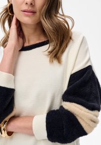 Gebreide trui met een crèmekleurig lijf, zwarte en beige pluizige mouwen, geribbelde manchetten en een ronde halslijn. Accent met een gouden manchetarmband.