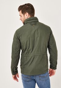 Olive green jacket with a zip closure, adjustable collar, and elastic cuffs. Features stitching accents and a logo patch at the back.