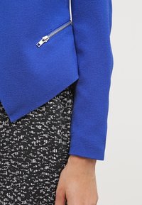 Modrá texturovaná bunda s diagonálním střihem a detaily na zip, nošená přes černé vzorované šaty.
