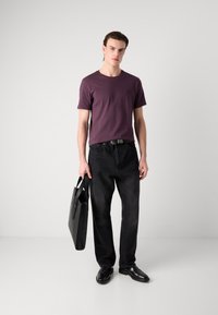 Tricou scurt lila cu un logo mic, blugi negri din denim și pantofi negri. Modelul ține o geantă elegantă neagră cu curea de umăr.