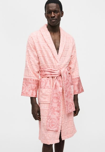 Άνδρας που φοράει ροζ υφασμάτινο μπουρνούζι με ανάγλυφα γράμματα και περίτεχνες μανσέτες με σχέδια, δεμένο στη μέση, στεκόμενος μπροστά σε απλό φόντο.