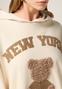 Hoodie beige clair avec "NEW YORK" en lettres brunes matelassées et un motif d'ours texturé. Tissu doux avec une coupe décontractée et une capuche.