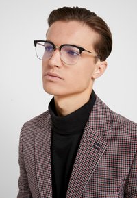 Ochelari negri și transparenți, de formă rectangulară, cu accente metalice, purtați cu un pulover negru și sub un sacou cu model roșu și alb.
