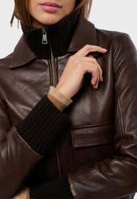 Braune Lederjacke mit einem gerippten schwarzen Kragen und braunen Strickbündchen. Sie verfügt über einen Frontreißverschluss und eine aufgesetzte Tasche auf der linken Seite.