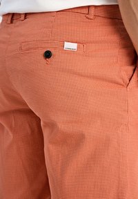 Pantaloni arancioni con texture, dotati di una piccola tasca posteriore con un bottone nero e un'etichetta bianca con il marchio sul lato.