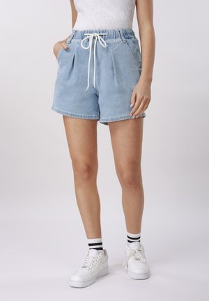 Donna che indossa pantaloncini di jeans azzurri chiari con coulisse bianca, sneakers bianche e calzini bianchi con strisce nere, in piedi con le mani nelle tasche.