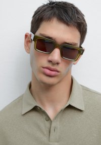 Polo shirt khaki e occhiali da sole rettangolari con montatura verde oliva e lenti scure, con un brand discreto sulla tempia.