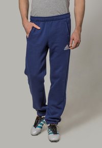 Mies, joka on pukeutunut tummansinisiin Adidas-housuihin ja valkoisiin kengtiin, joissa on teal-vihreät raikui, seisoo toinen käsi taskussa harmaata taustaa vasten.