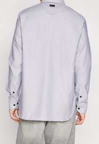 Vaaleanharmaa pitkähiha-paita, jossa nappikiinnitys kauluksessa, mustat koristeraidat kalvosimissa ja selässä olkapääntuki. Sileä kangaspinta.